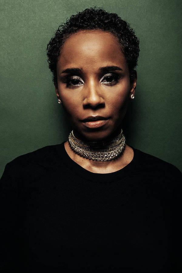 Portrait of a stylish adult woman with short curly hair wearing a black shirt and layered silver choker against a green backdrop.