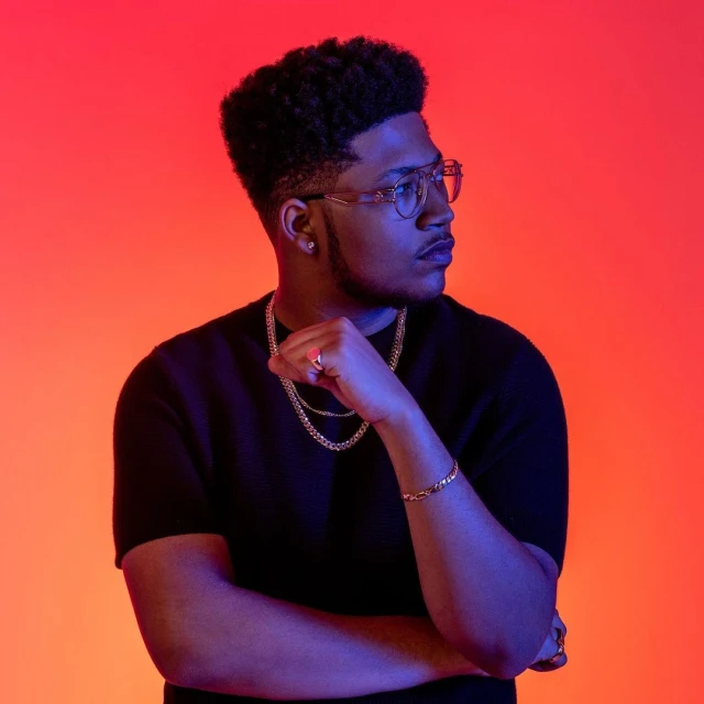 Portrait of a stylish young adult male with dark curly hair and glasses, wearing a black top and gold jewelry, against a colorful backdrop.