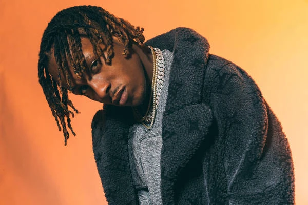 Close-up studio shot of a stylish African-American male model wearing a gray coat and a chain posing against a warm orange background.