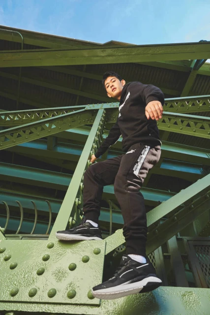 Man in black sweatsuit poses on a green metal structure against a blue sky, showcasing a Puma branded athletic outfit and sneakers.