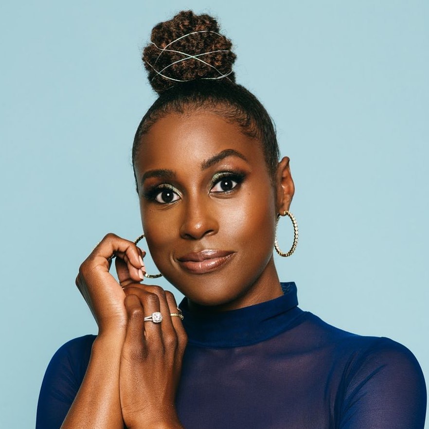Close-up of an adult female with a hair bun, wearing a sheer blue top, gold hoop earrings, and a diamond ring against a light blue background.