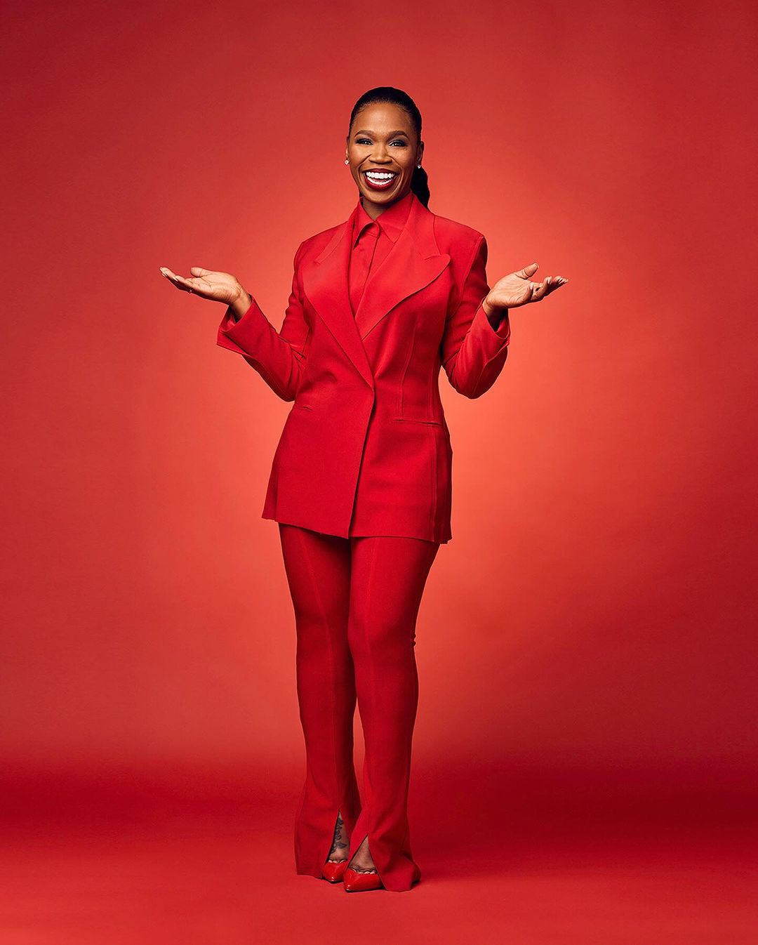 A full studio portrait of a smiling Black woman in a red pantsuit, with an open hand gesture, set against a red background.