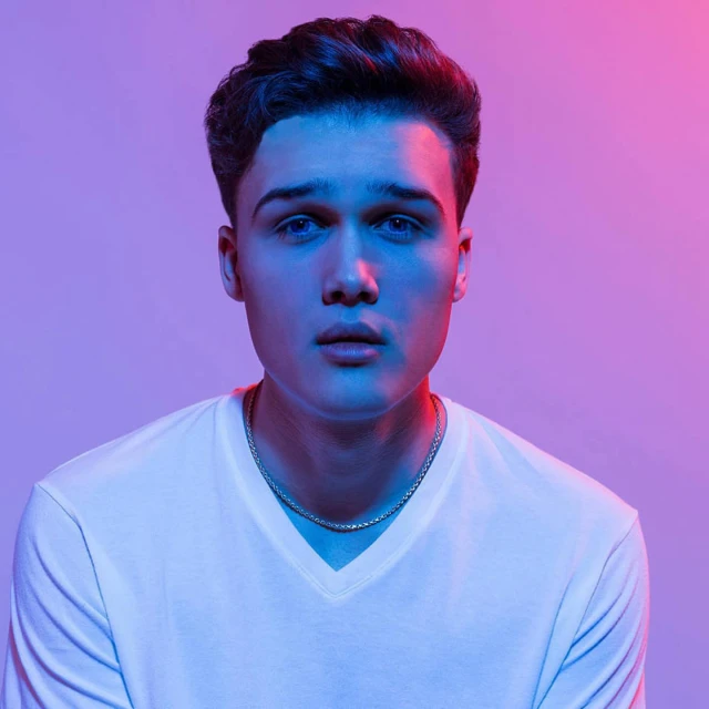 Portrait of a young adult male with dark hair and blue eyes, wearing a white t-shirt, illuminated by blue and red lighting.