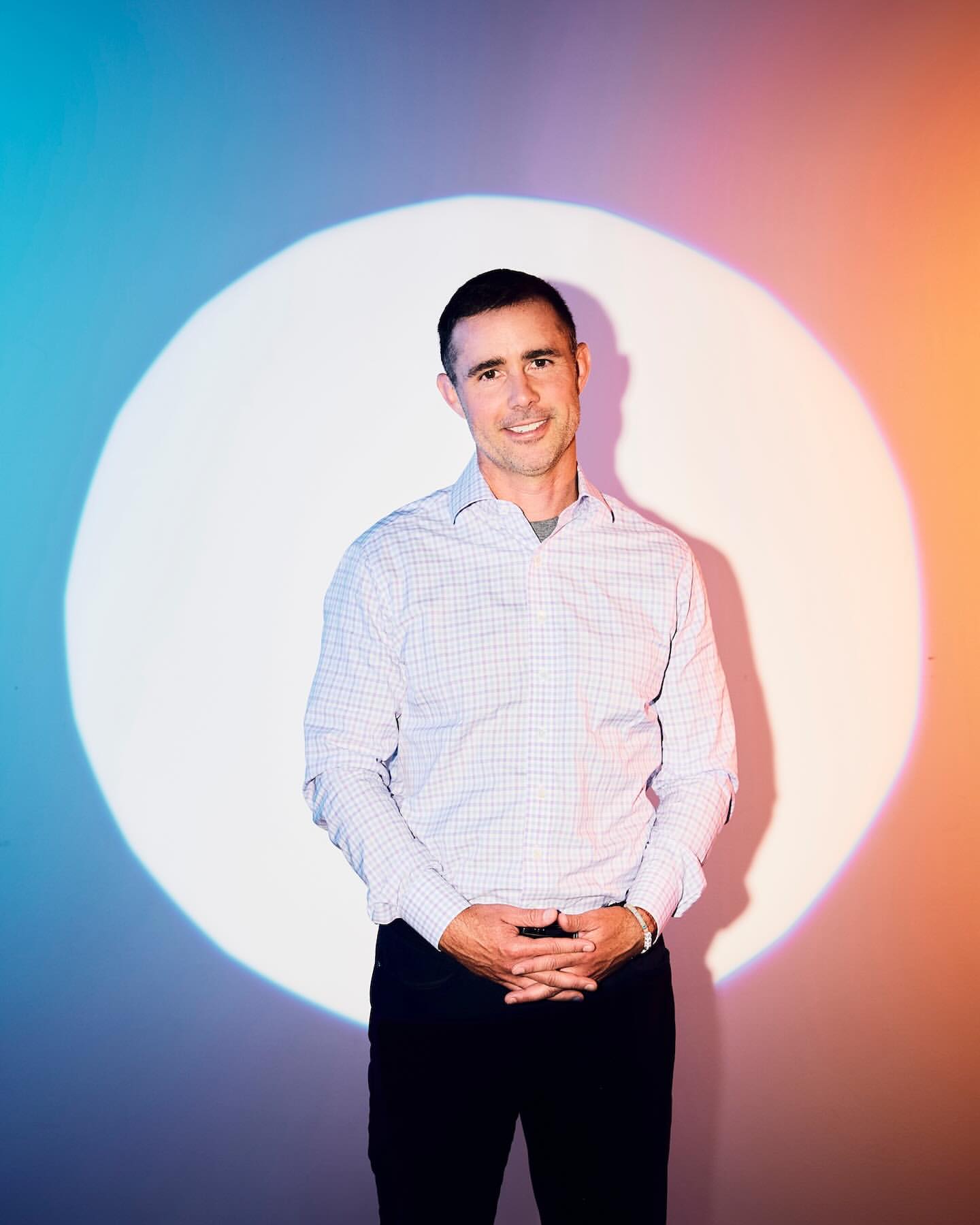 Adult male with short dark hair poses in front of white wall with bright spot lighting and blue and orange gradient color.