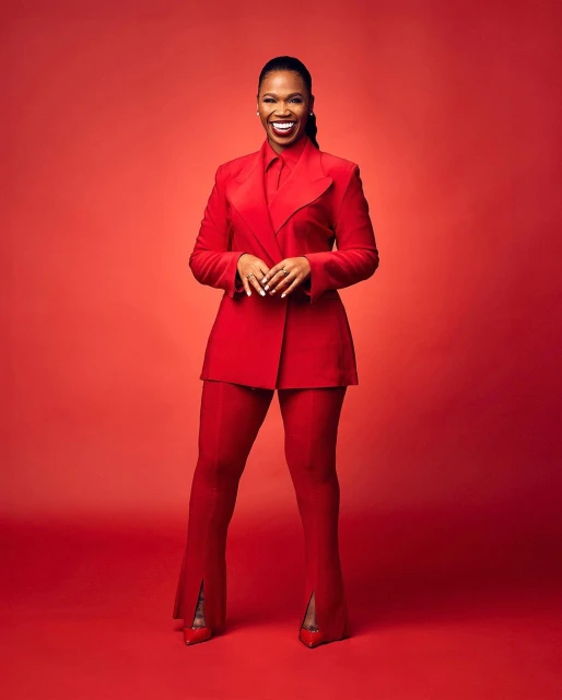 A confident, middle-aged African-American woman in a red suit stands against a red backdrop, smiling with hands clasped at her waist.