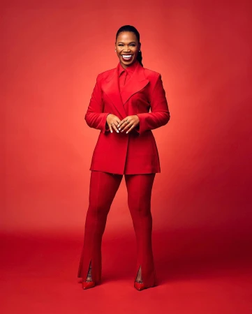 A confident, middle-aged African-American woman in a red suit stands against a red backdrop, smiling with hands clasped at her waist.