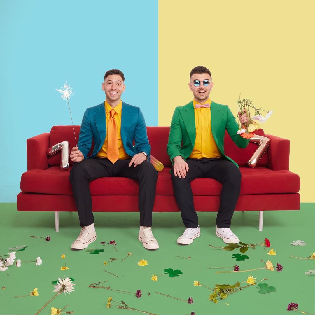 Two stylish men sit on a red couch in front of a split blue and yellow background, celebrating with flowers and a sparkler.