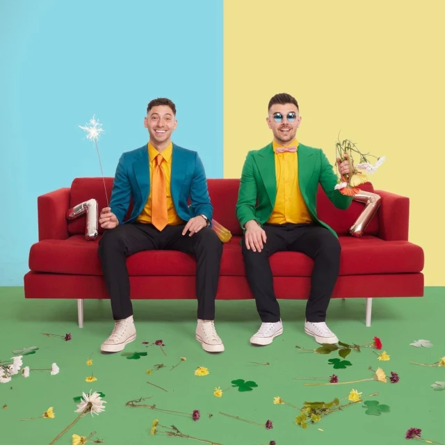 Two stylish men sit on a red couch in front of a split blue and yellow background, celebrating with flowers and a sparkler.