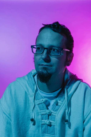 Close-up portrait of an adult man wearing glasses and a hooded shirt, set against a gradient background of pink and blue.