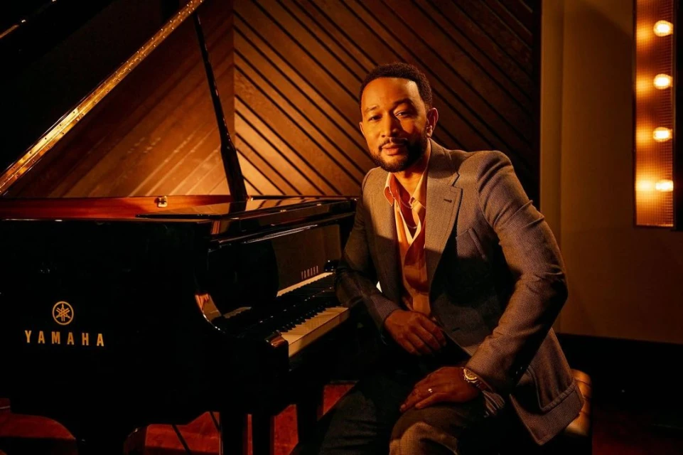 John Legend poses beside a piano in a photo studio with wood paneled walls and accent lighting, creating a sophisticated and warm ambiance.