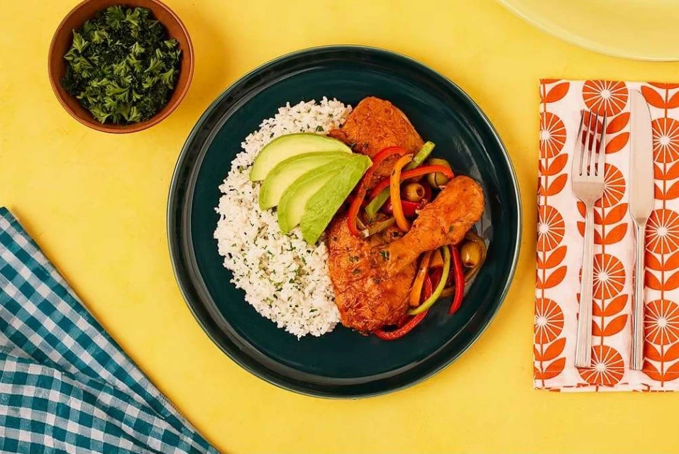 Overhead shot of appetizing chicken and rice dish garnished with avocado and red and green bell peppers, served on a dark green plate.