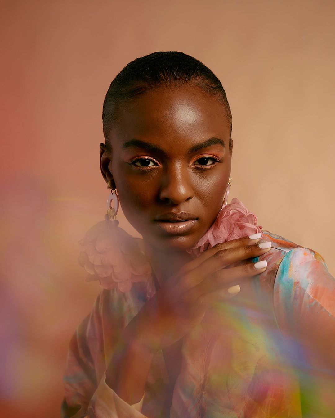 Portrait of a Black woman with short hair wearing a colourful blouse, large earrings and pink eye makeup against a tan backdrop.