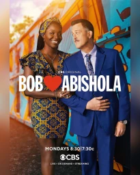 Man and woman standing together smiling, the woman in a patterned blue and yellow dress and the man in a blue suit.