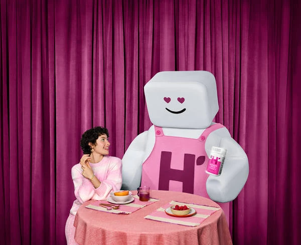 Woman in pink striped sweater seated at a round table with a large mascot figure holding a supplement bottle