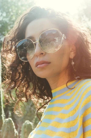 Close-up portrait of a woman wearing oversized sunglasses and a yellow and blue striped sweater outdoors.