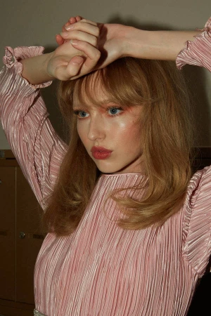 Woman with blonde hair and pink eyeshadow wearing a pleated pink dress with her hands clasped above her head.