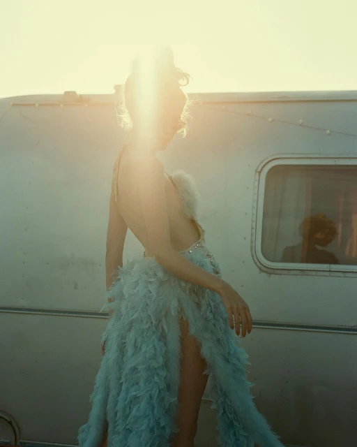 Adult female in feathered dress poses next to a vintage travel trailer at golden hour, bathed in warm sunlight with lens flare.