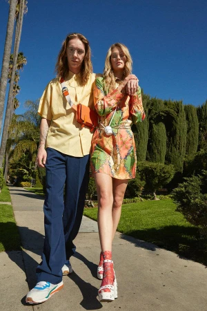 Two models walking side by side on a sunny sidewalk lined with trees under a clear blue sky