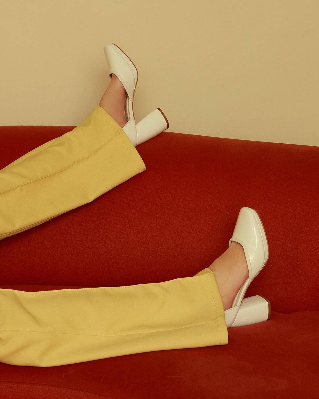 Fashionable leg display featuring white high heel mules, mustard yellow flared trousers, and a red textured surface backdrop.