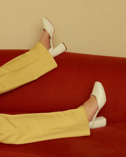 Fashionable leg display featuring white high heel mules, mustard yellow flared trousers, and a red textured surface backdrop.