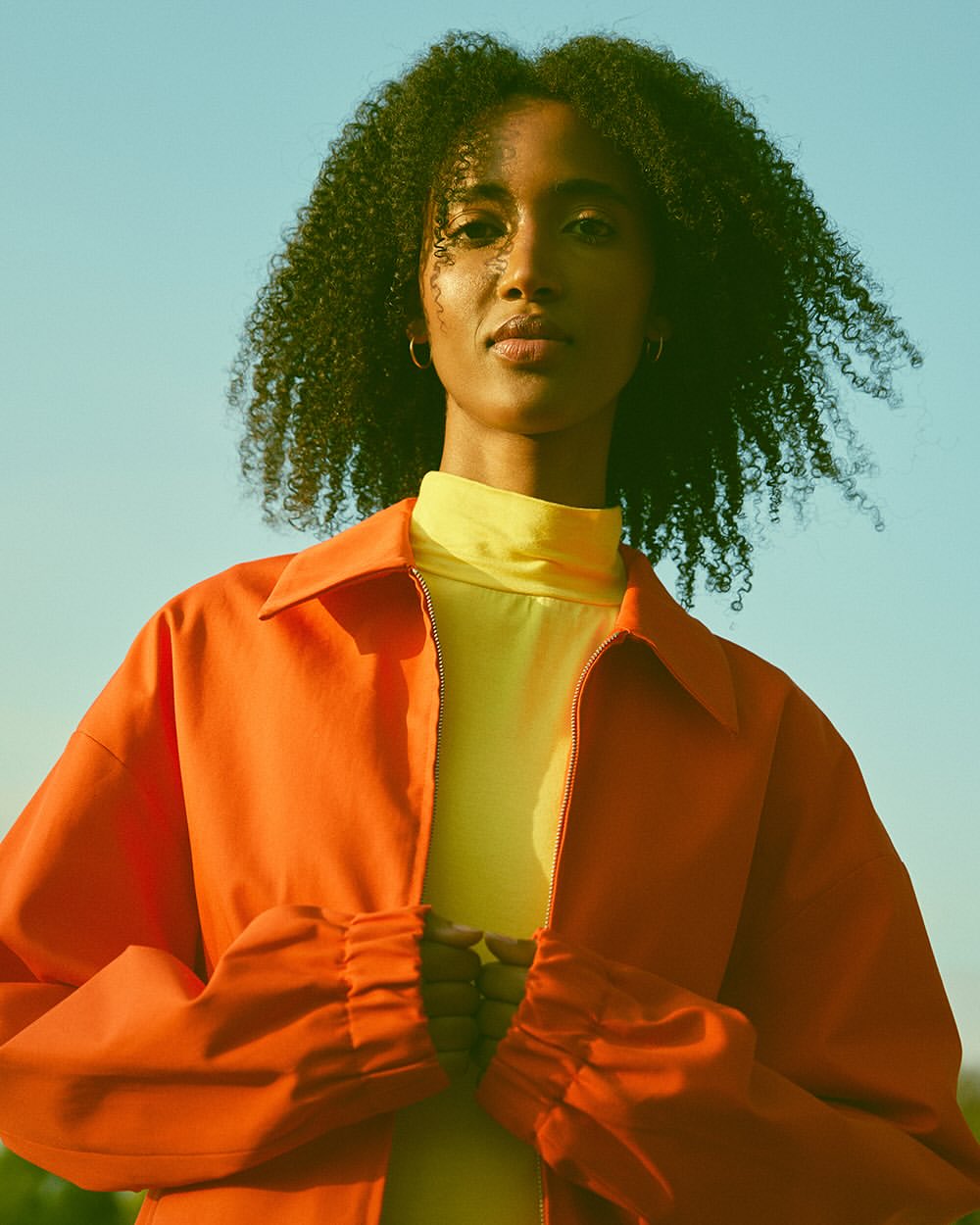 Close-up studio shot of an adult woman with dark, curly hair wearing an orange jacket and yellow top against a clear sky backdrop.