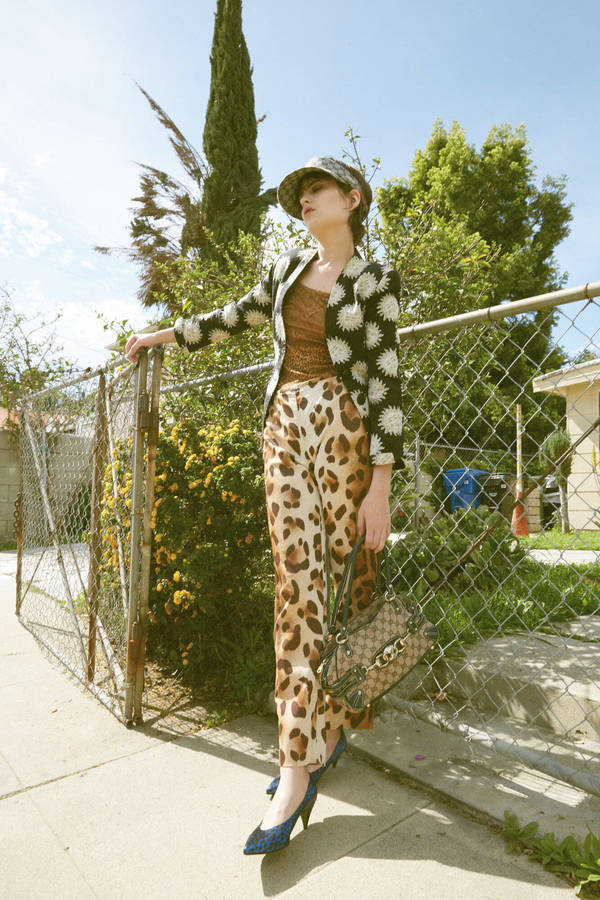 Woman in patterned jacket and animal print pants stands outdoors with handbag and heels against a fence, stylishly posing in daylight.