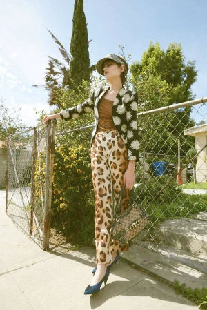 Woman in patterned jacket and animal print pants stands outdoors with handbag and heels against a fence, stylishly posing in daylight.