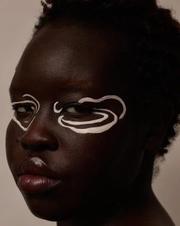 Close-up portrait of a Black woman with short dark hair and white abstract face paint around her eyes.