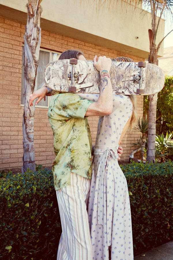 A man and woman embracing outdoors while holding up a skateboard that obscures their faces, photographed by Katy Pritchett.