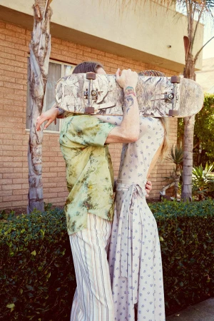 A man and woman embracing outdoors while holding up a skateboard that obscures their faces, photographed by Katy Pritchett.