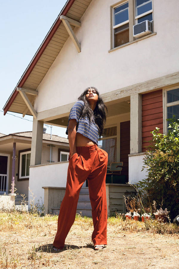 Adult woman with dark hair poses outdoors in front of white house wearing blue and white striped crop top and rust-colored pants.