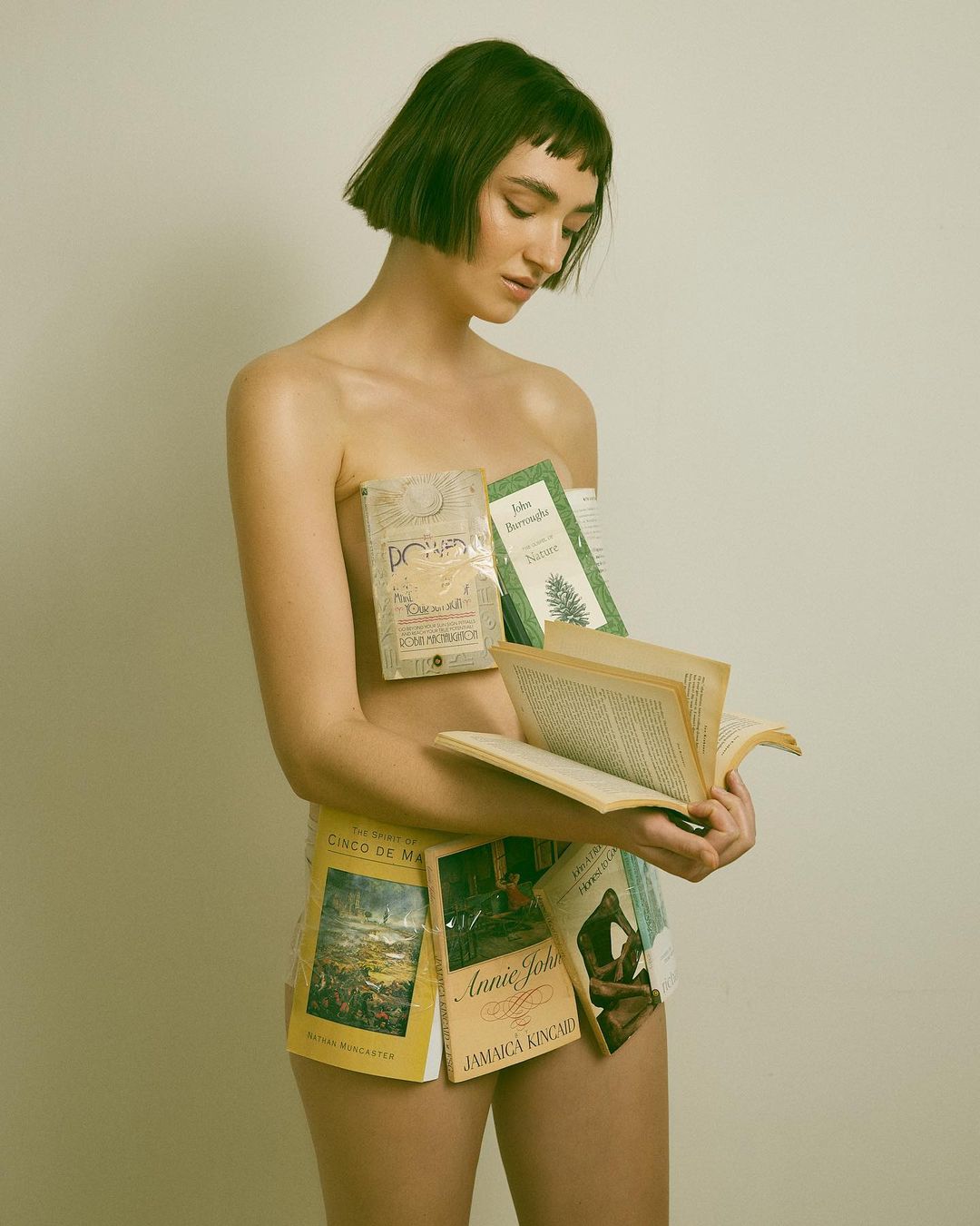 Young woman stands indoors, partially covered with books, glancing downward while holding an open book in her hands against a beige backdrop.