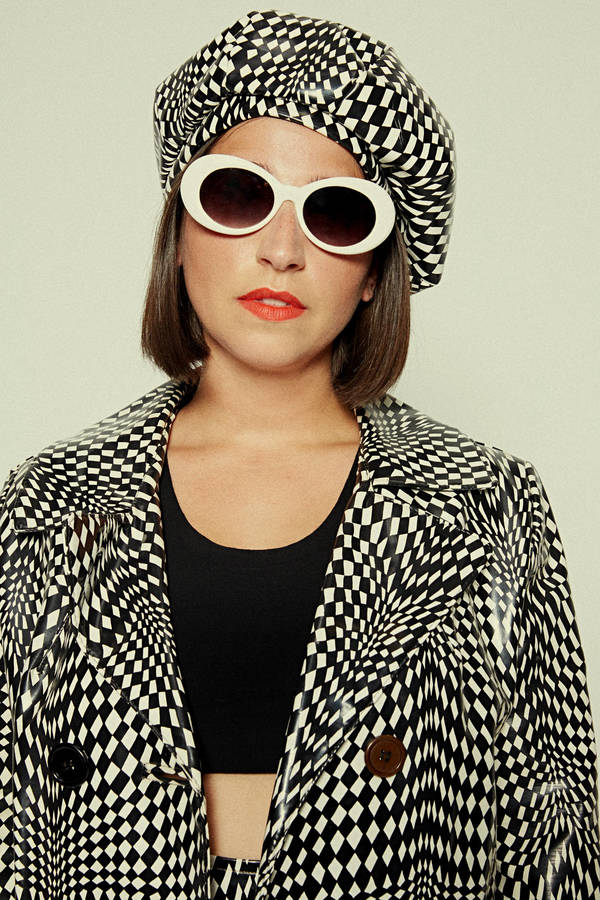 Woman in a patterned black and white coat and beret posing with white sunglasses and red lipstick against a neutral backdrop.