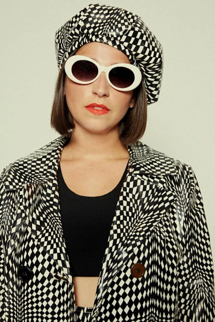 Woman in a patterned black and white coat and beret posing with white sunglasses and red lipstick against a neutral backdrop.