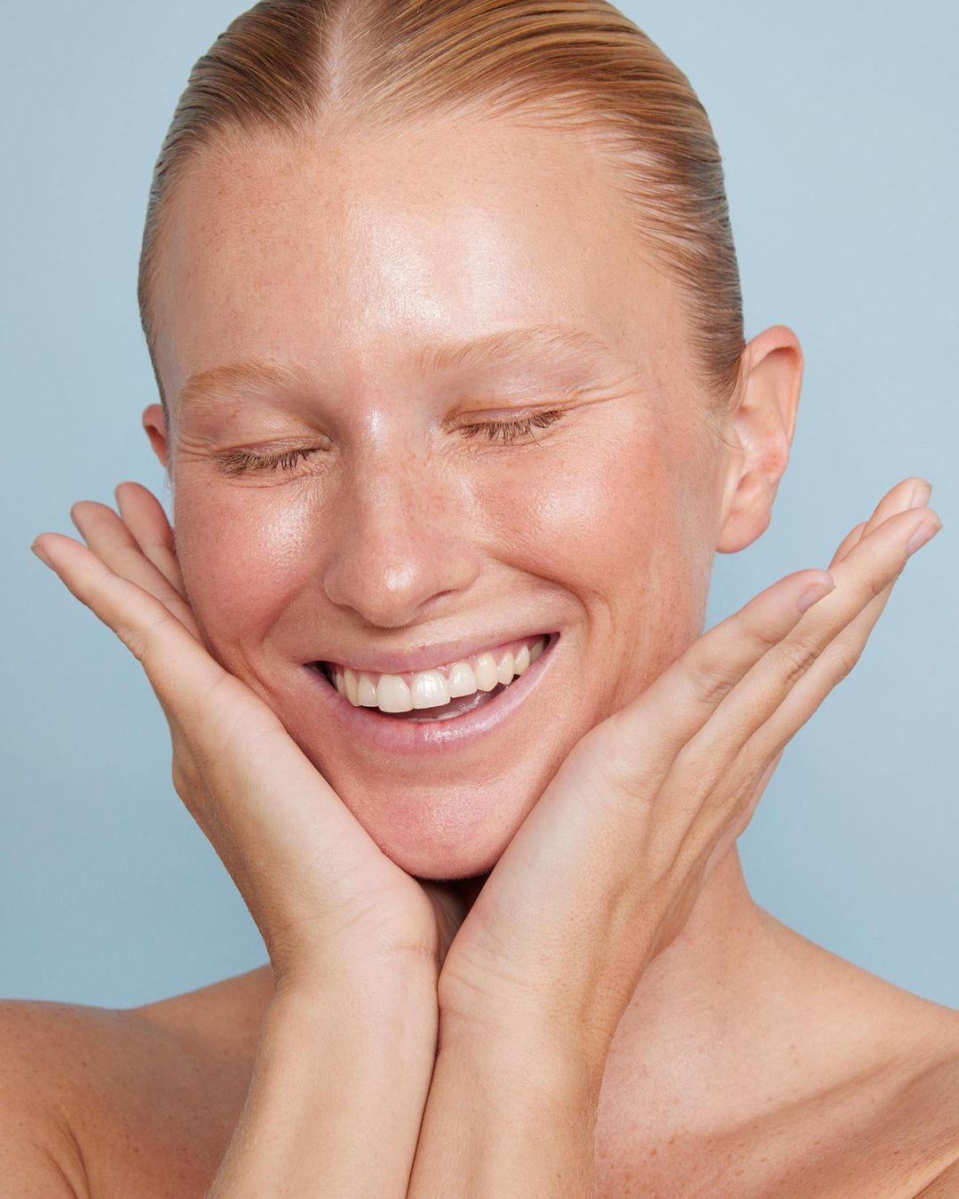 Close-up shot of a smiling fair-skinned woman with hands on her cheeks, eyes closed, showing a serene expression against light blue background.