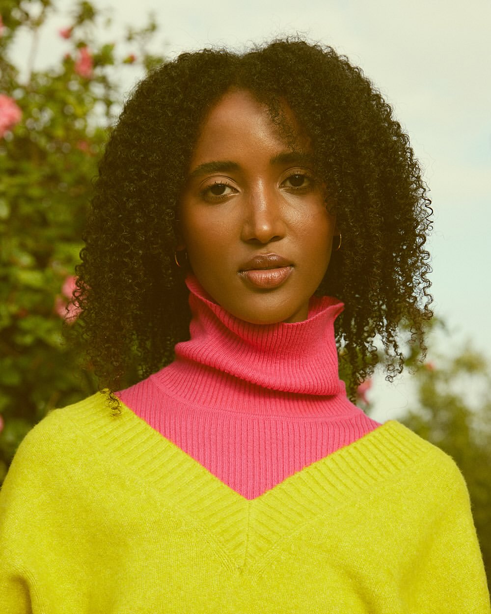 A young woman with curly hair poses outdoors wearing a yellow and pink sweater, with flowering bushes softly blurred in the background.