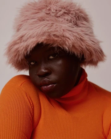 Black woman wearing a fluffy pink bucket hat and an orange ribbed turtleneck sweater against a pale background