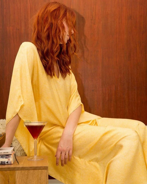 Woman in yellow dress sits with cocktail, cassette player, and books against a wood wall. Red hair cascades down her shoulder.