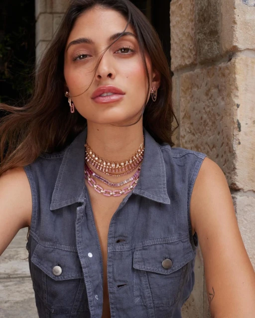 Brunette woman poses with layered necklaces and a sleeveless denim jacket against a stone wall in a close-up portrait shot.