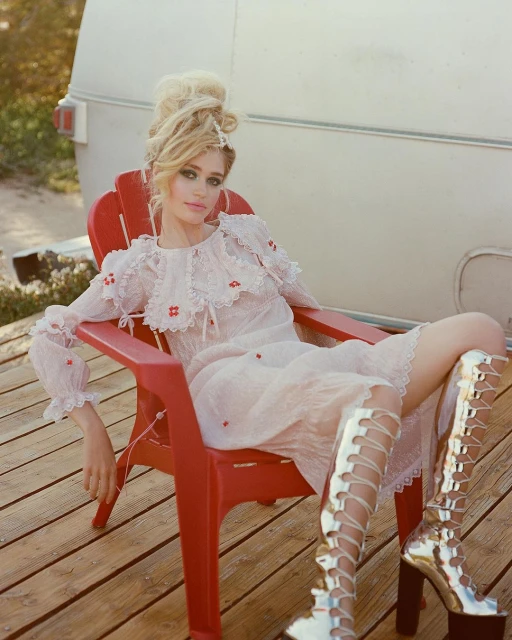 A young adult woman in a lacy dress poses in a red Adirondack chair on a wooden deck, with a vintage camper in the background.