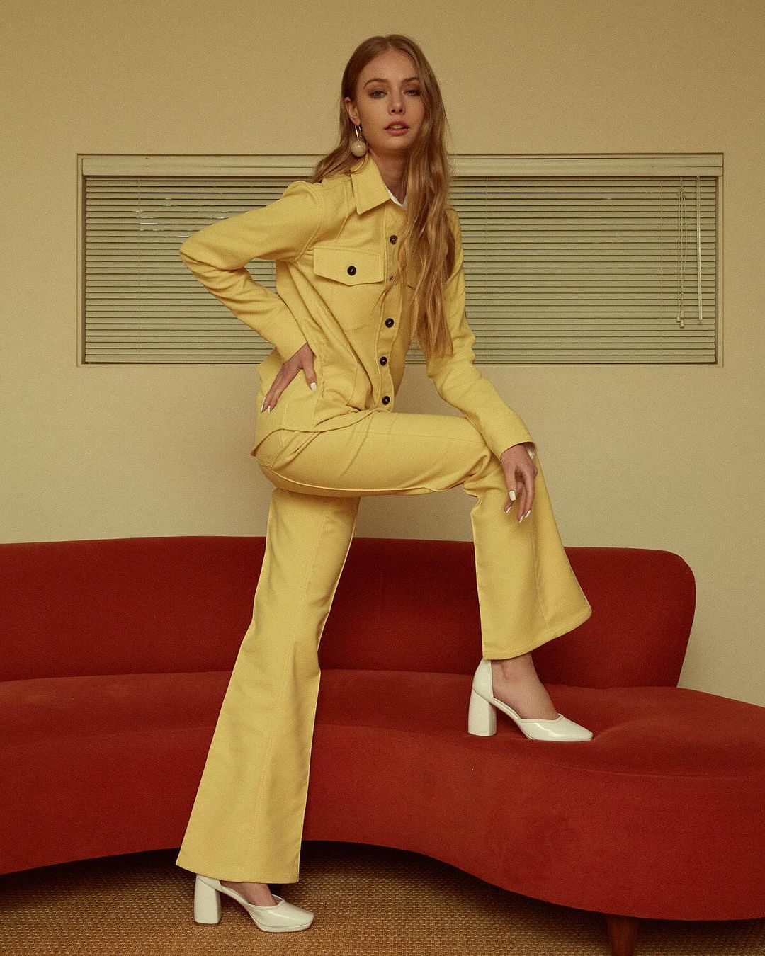 Model in a yellow button-up shirt and matching flared trousers standing with one foot on a red curved sofa.