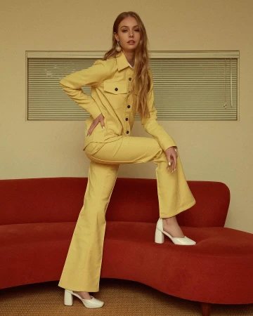Model in a yellow button-up shirt and matching flared trousers standing with one foot on a red curved sofa.