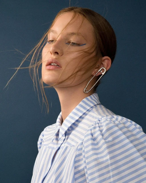 A woman poses in a blue and white striped shirt, wearing a safety pin earring, against a dark blue background, with loose hair.
