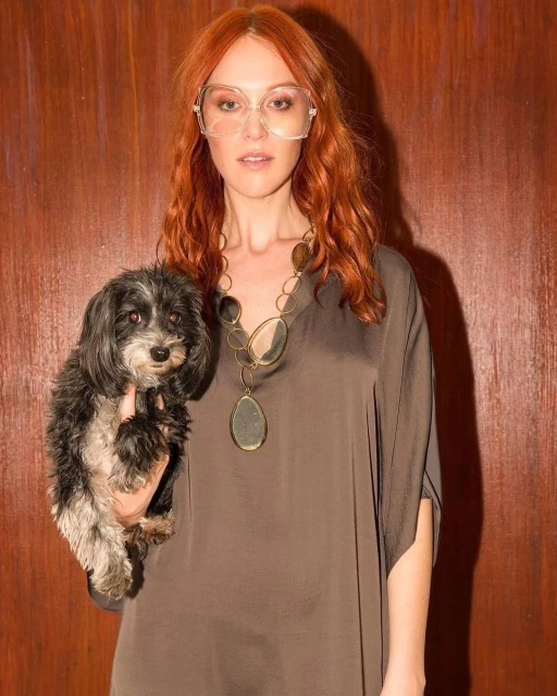 An adult woman with red hair stands indoors holding a small black dog, wearing eyeglasses and a necklace with dark wood-paneled wall behind.