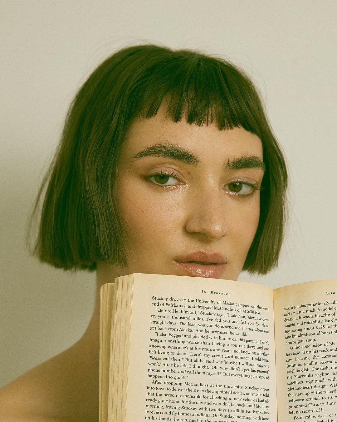 Portrait of a young woman with a blunt bob haircut holding a book up to her chest against a neutral background in a studio setting.