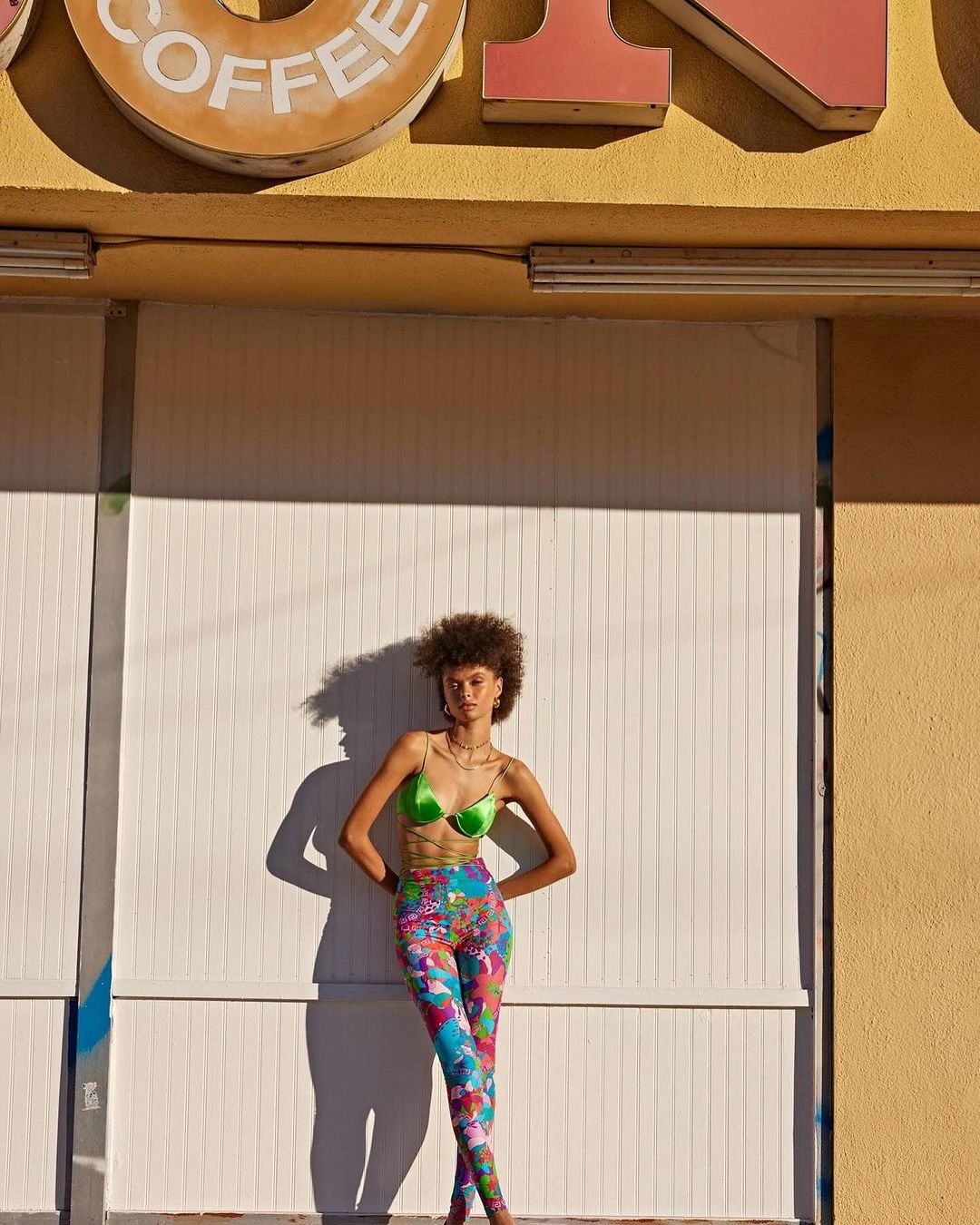 Adult woman with curly hair poses in front of a closed coffee shop in a green bra top and patterned leggings in an outdoor setting.