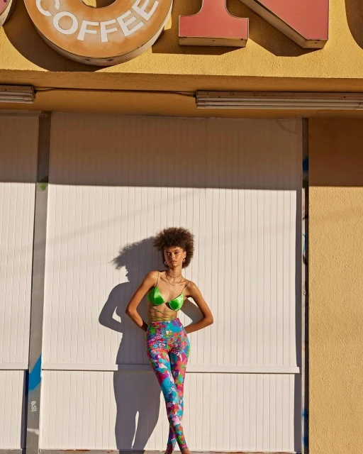 Adult woman with curly hair poses in front of a closed coffee shop in a green bra top and patterned leggings in an outdoor setting.