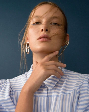 Portrait of a woman wearing a blue and white striped shirt and silver safety pin earrings against a dark blue background.