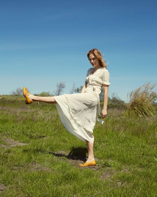 Young woman in a cream-colored dress playfully poses with a raised leg in a sunny field, showcasing yellow leather shoes against a blue sky.
