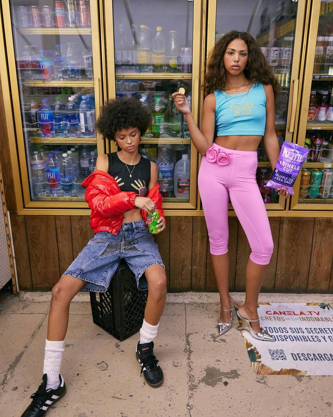 Two stylish women pose indoors near glass refrigerators. One sits on a crate, holding a soda, while the other holds chips. 